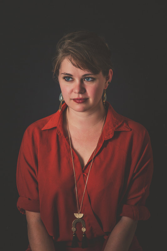 The poet Molly Bess Rector is shown from the waist up. She is wearing an orange collared shirt with sleeves rolled up, a long gold gold necklace, and dark lipstick. Her hair is pulled back and she looks into the distance.
